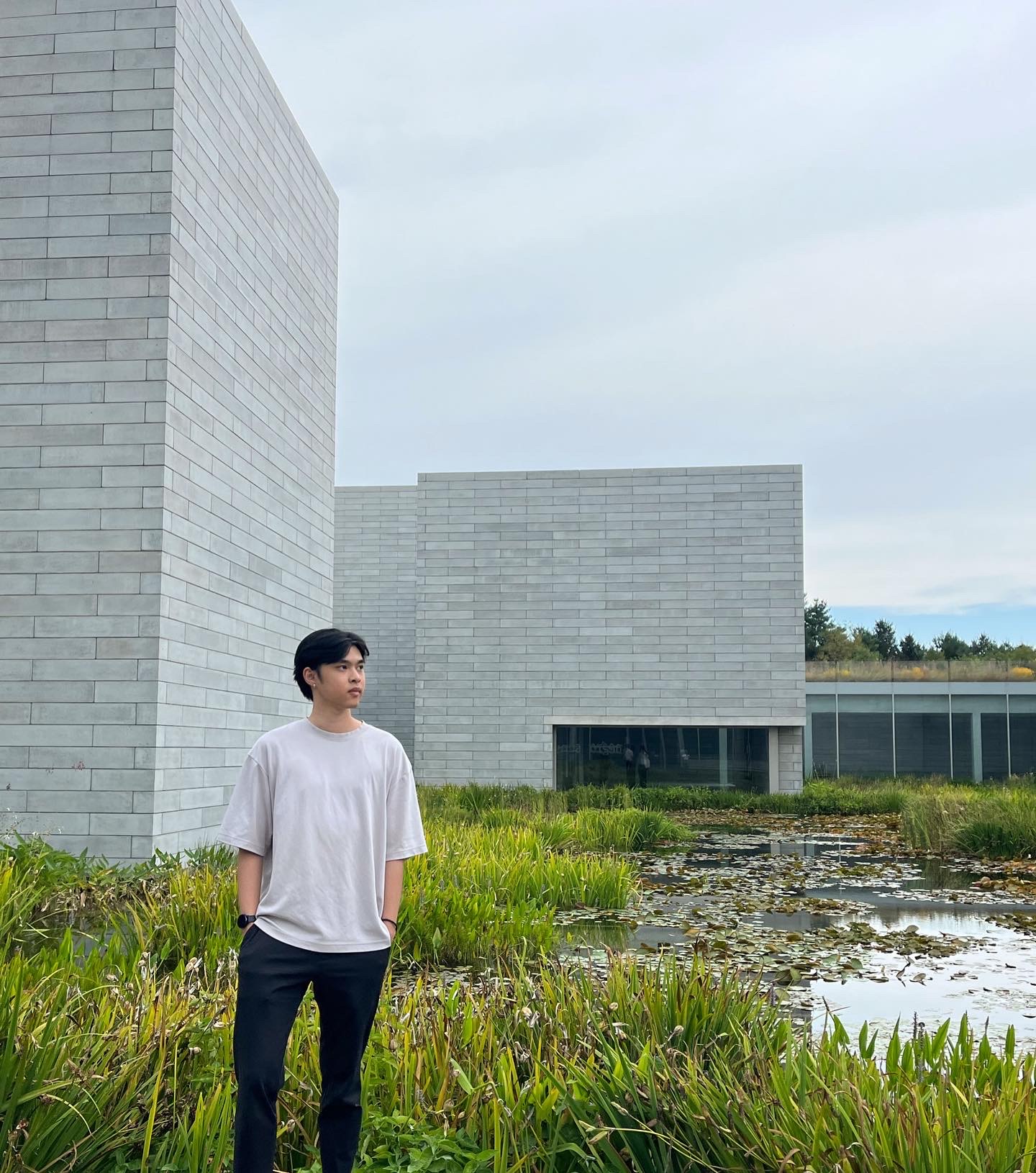 Young man in a white shirt and black pants standing in front of tall green plants and a pond with lily pads beside modern gray brick buildings under a cloudy sky.