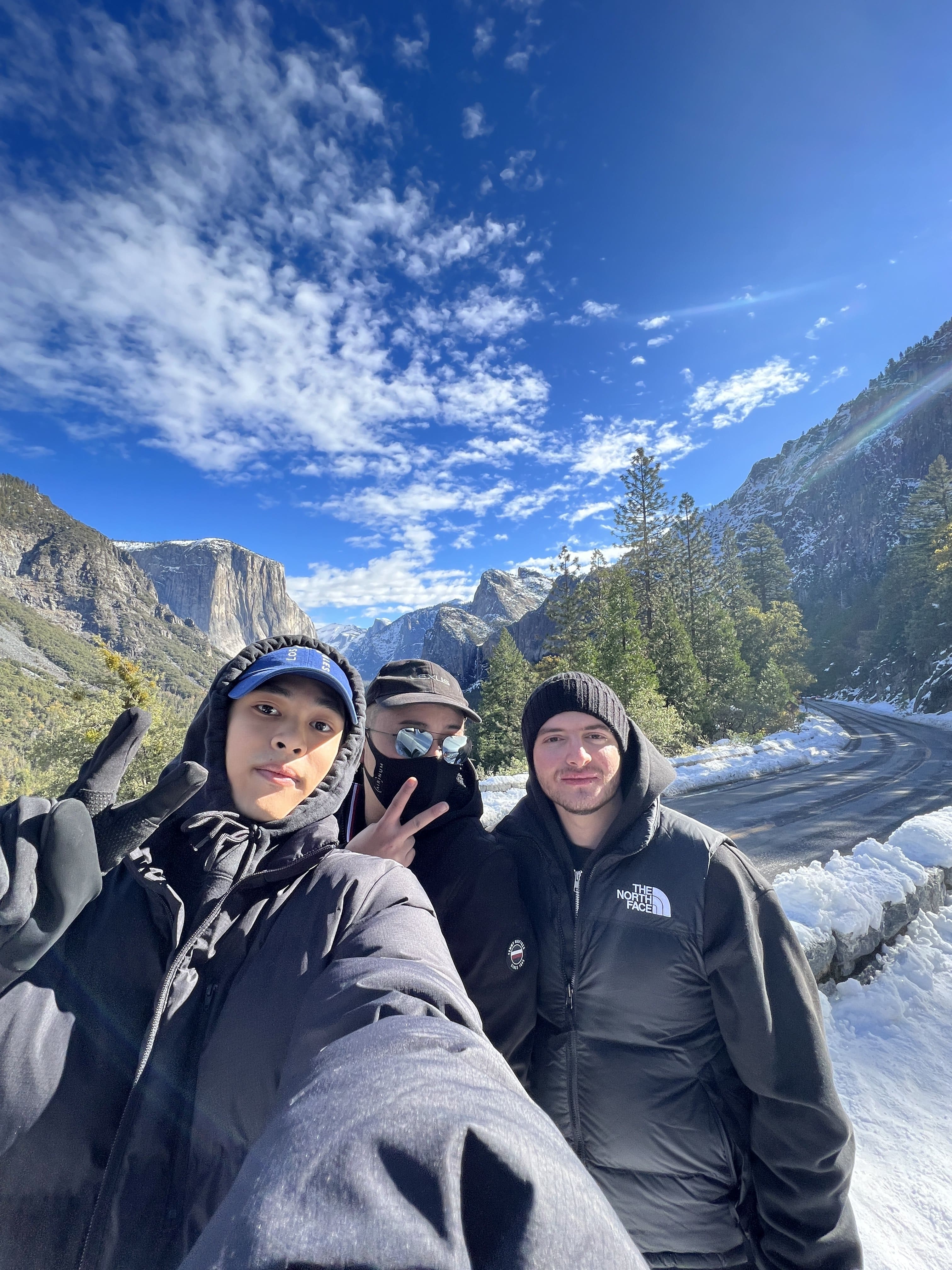 Three young men dressed in winter jackets taking a selfie on a snowy mountain road with pine trees and rocky cliffs under a bright blue sky.